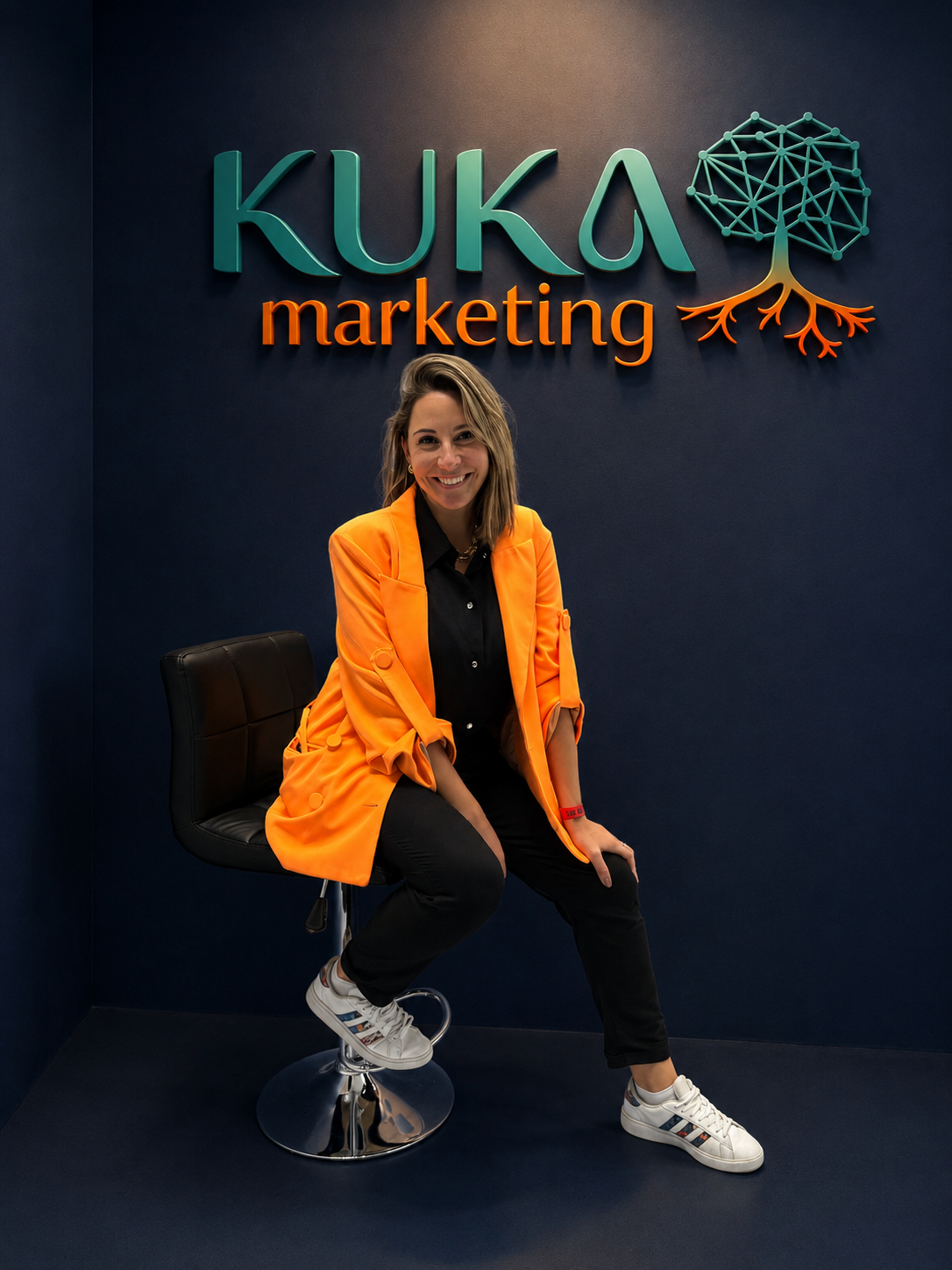 Aline, a Kuka, sentada no escritório da KUKA Marketing com o logo da marca na parede